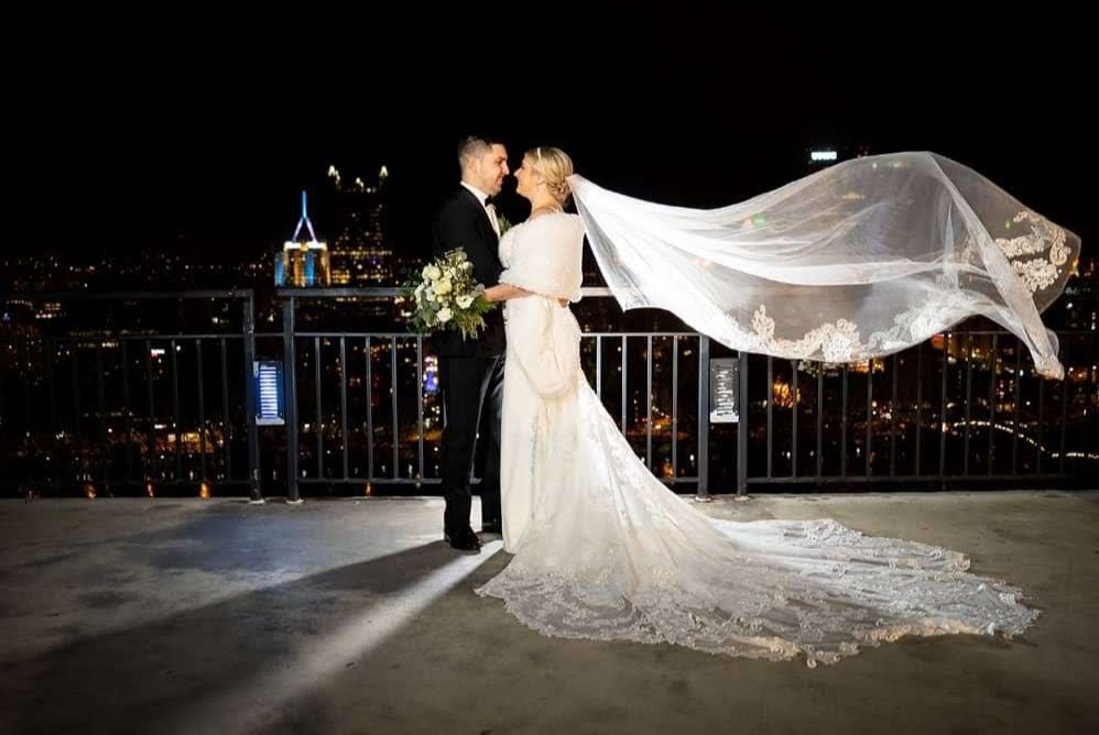 Bride and groom rooftop portrait with veil flowing against city skyline