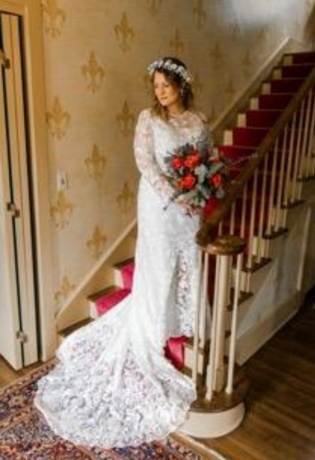 Bride in long-sleeve lace gown on staircase with red bouquet